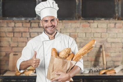 smiling young male baker holding loaf of breads in paper bag showing thumb up sign